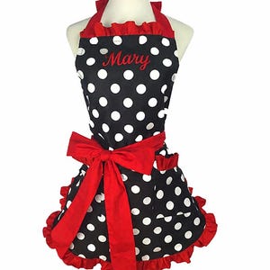 May include: A black apron featuring a white polka dot pattern, accented with red ruffles along the neckline and hem. The apron includes a large red waist tie and two pockets, with the name Mary embroidered in red script on the upper chest area.