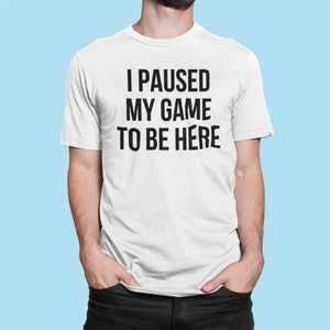 May include: White t-shirt with the text "I PAUSED MY GAME TO BE HERE" in bold black letters. The shirt is being worn by a person with their hands in their pockets. The background is a light blue.