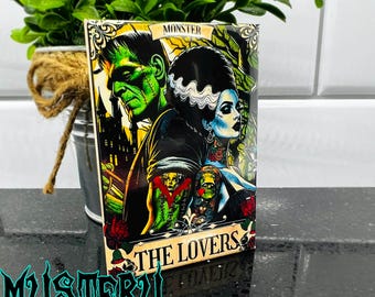 Puede incluir: Una calcomanía rectangular con una ilustración gótica de Frankenstein y su novia, con el texto "THE LOVERS". La imagen tiene una paleta de colores oscuros con acentos verdes, negros y rojos. La palabra "MONSTER" está en la parte superior.