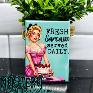 May include: A turquoise blue square magnet with a vintage-style illustration of a woman in a pink dress holding a strawberry cake. The text on the magnet reads "Fresh Sarcasm served DAILY."