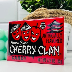May include: A red and white candy wrapper with three cartoon cherries with menacing expressions. The wrapper says "Ferrara Pan Cherry Clan Candy" and "Net Wt 7/8 oz 25g".