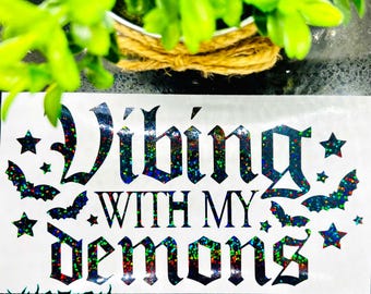 May include: A black and white vinyl sticker with a glittery rainbow finish. The sticker says "Vibing with my demons" in a gothic font. There are stars and bats around the text.