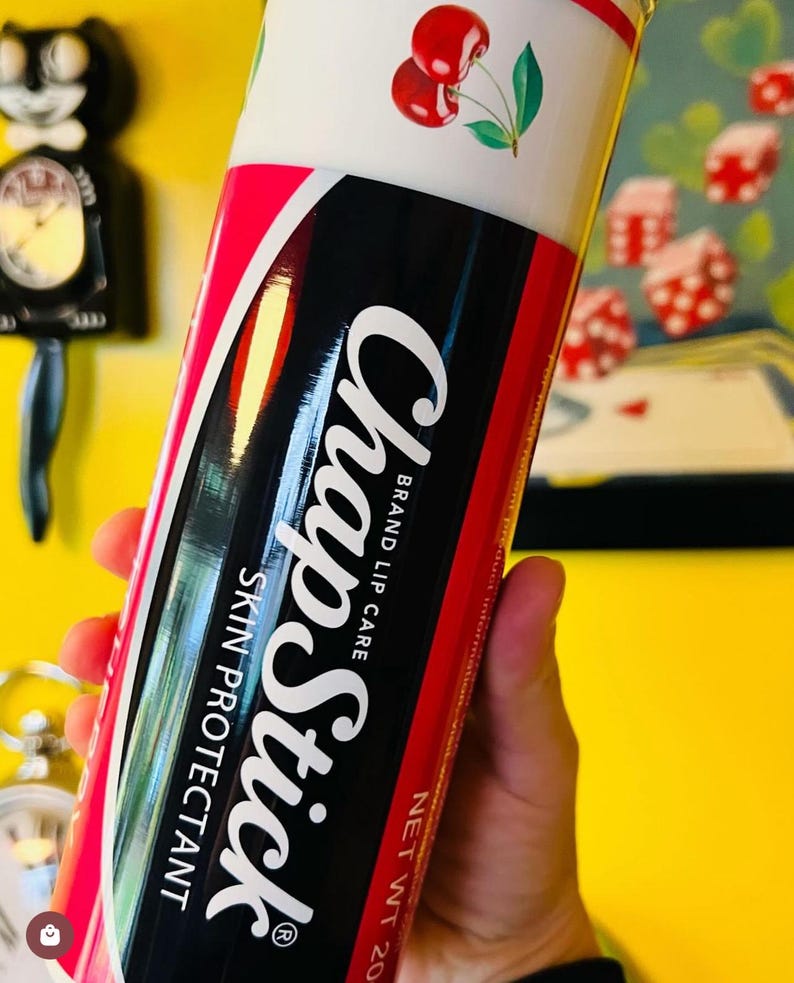Pu&ograve; includere: Un tubo rosso e nero di balsamo per labbra ChapStick con un disegno di ciliegie sulla parte superiore. Il tubo riporta la scritta "ChapStick Brand Lip Care Skin Protectant" e "Net Wt. 20 oz."