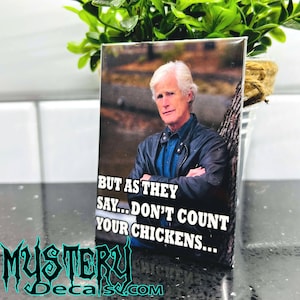 May include: A white magnet with a photo of a man with gray hair and a black jacket. The text on the magnet reads "But as they say... don't count your chickens..."