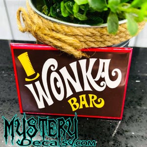 May include: A brown and red metal sign with white lettering that reads "Wonka Bar". A yellow top hat is above the word "Wonka".