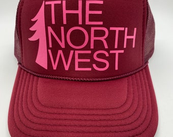May include: A burgundy trucker hat with a pink embroidered logo that says "THE NORTH WEST" and a small pine tree graphic.