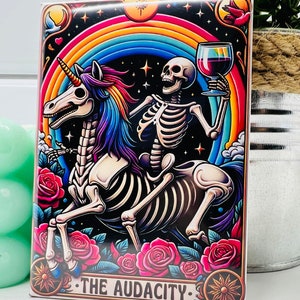 May include: A colorful illustration of a skeleton riding a unicorn with a rainbow in the background. The skeleton is holding a glass of red wine. The text "THE AUDACITY" is at the bottom of the image.