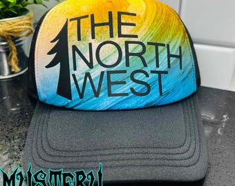 May include: A black trucker hat with a gray brim and a colorful top panel. The top panel features a design with the words "THE NORTH WEST" in black, along with a black tree graphic. The background has a gradient of yellow, blue, and green.