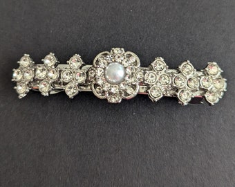 May include: Silver-toned barrette with a central flower design featuring a pearl-like bead, surrounded by sparkling clear rhinestones. The barrette has floral clusters on each side, creating a decorative hair accessory.
