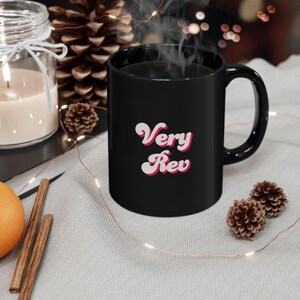 Pode incluir: Uma caneca de cerâmica preta com as palavras "Very Rev" em uma fonte retrô. A caneca está cheia de um líquido escuro, possivelmente café ou chá, e está fumegando. A caneca está em uma superfície cinza claro com elementos decorativos.