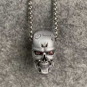 Könnte beinhalten: Eine silberfarbene Halskette mit einem detaillierten Totenkopf-Anhänger mit roten Augen. Der Totenkopf ist eine Darstellung eines Roboters oder Cyborgs mit aufwendigen Details und einem mechanischen Design.