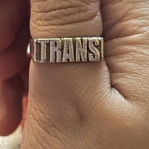 Peut inclure: Une bague en argent avec le mot "TRANS" gravé dessus.