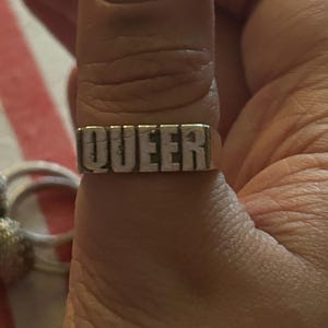May include: A silver ring with the word "QUEER" engraved in white lettering.