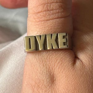 Può includere: Un anello d'argento con la parola "DYKE" incisa in lettere dorate.