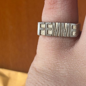 May include: A silver ring with the word "FEMME" in raised capital letters. The ring is worn on a finger, with the word "FEMME" across the top. The ring has a textured finish, possibly brushed. The ring is a statement piece.