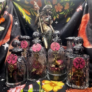 May include: An assortment of decorative glass bottles with black wax accents, each adorned with a pink wooden tag and a gold key charm. The bottles are filled with dried flowers. A bronze statue and floral fabric are in the background.