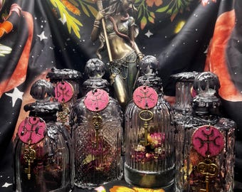 May include: An assortment of decorative glass bottles with black wax accents, each adorned with a pink wooden tag and a gold key charm. The bottles are filled with dried flowers. A bronze statue and floral fabric are in the background.