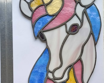 Può includere: Un acchiappasole in vetro colorato a forma di unicorno con segmenti di vetro bianco, giallo, rosa e blu uniti da linee di piombo scure. Un righello metallico a sinistra indica un'altezza totale di circa 25,4 cm.