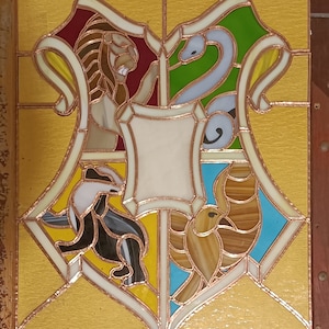 Può includere: Una finestra in vetro colorato che raffigura gli stemmi delle case di Hogwarts. Gli stemmi hanno la forma di uno scudo con un leone, un'aquila, un tasso e un serpente. Il leone è rosso, l'aquila è gialla, il tasso è nero e il serpente è verde.