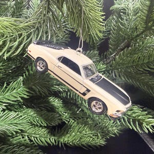 May include: A cream-colored classic car ornament with a black stripe on the hood and roof. The ornament is hanging from a silver wire against a backdrop of green Christmas tree branches. The car has silver wheels.
