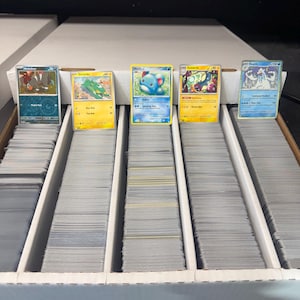 May include: A collection of Pokémon trading cards, featuring various characters and designs. The cards are displayed in a divided storage box, with several stacks of cards visible. The cards have different colors and illustrations, with text and numbers.