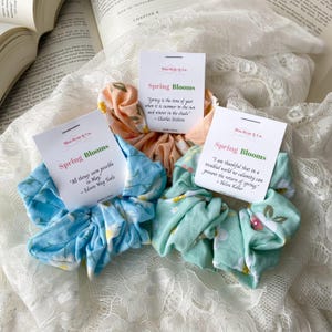 Puede incluir: Tres scrunchies de pelo de tela con estampados florales. Los scrunchies son en tonos azul, verde y melocotón. Cada scrunchie tiene una pequeña tarjeta adjunta con una cita sobre la primavera. Las citas son: "Todo parece posible en mayo" - Edwin Way Teale, "La primavera es la época del año en que hace verano al sol e invierno a la sombra" - Charles Dickens, y "Estoy agradecida de que en un mundo problemático ninguna calamidad pueda impedir el regreso de la primavera" - Helen Keller.