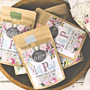 May include: Several kraft paper packets of "Sweet Pickins Milk Paint" are arranged in a wooden bowl. The packets feature floral designs and color swatches in shades of blue and green. The text "bluebird" and "oh olive!" are visible.