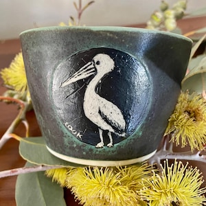 May include: A dark green ceramic bowl featuring a circular inset with a white, hand-painted illustration of a pelican. The bird is depicted in profile, and the word "cook" is written in small white letters near the base of the circular design.