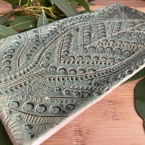 May include: A rectangular, handmade ceramic tray with a leaf design. The tray is a muted teal colour with intricate patterns carved into the surface. The leaf design is centered, with detailed lines and circles. The tray is surrounded by eucalyptus leaves.