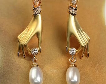 Puede incluir: Un par de pendientes dorados con diseño de mano. Cada pendiente tiene una mano que sostiene una gema redonda y brillante, con una perla en forma de lágrima colgante. Los pendientes tienen un acabado detallado y texturizado.