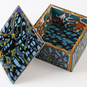 Könnte beinhalten: Geöffnete quadratische Box mit Deckel, mit einem farbenfrohen abstrakten Design. Deckel und Außenseite haben eine schwarze Basis mit blauen, grünen, gelben und violetten Mustern. Der Innenraum hat ein blaues und orangefarbenes Design.