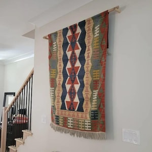 May include: A rectangular woven wall hanging featuring a geometric pattern in shades of rust orange, navy blue, olive green, and cream. The textile is suspended from a light-colored wooden rod and finished with a fringe along the bottom edge.
