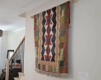 May include: A rectangular woven wall hanging featuring a geometric pattern in shades of rust orange, navy blue, olive green, and cream. The textile is suspended from a light-coloured wooden rod and finished with a fringe along the bottom edge.