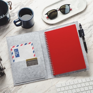 May include: A flat lay featuring a red spiral notebook, gray felt folio, sunglasses, a coffee mug, and a vintage camera. An open folio displays a red notebook and a pocket with an Air Mail envelope and a card. A keyboard and a book are also visible.