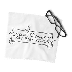 May include: A white microfiber cleaning cloth with the text "good moms SAY BAD WORDS" in black script and a heart symbol. Black-framed eyeglasses rest on the cloth, which has a scalloped edge.