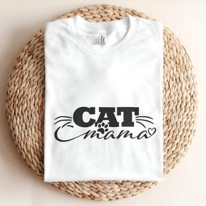 以下が含まれることがあります： 前面に黒い文字でCAT mamaとプリントされた白い半袖Tシャツ。デザインには、mamaという単語の上に小さな肉球のマーク、CATという単語から伸びる様式化されたヒゲ、そして最後に小さなハートが含まれています。
