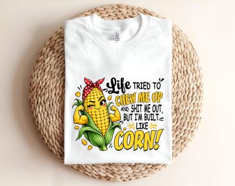 May include: A white t-shirt featuring a graphic of a winking corn cob character wearing a red polka dot headband and flexing its arms. The text reads: Life TRIED TO CHEW ME UP AND SHIT ME OUT, BUT I'M BUILT LIKE CORN!