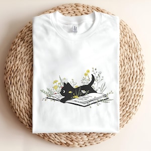 以下が含まれることがあります： 白い半袖Tシャツ。開いた本の上に横たわる黒猫と、その周りに描かれた黄色、白、緑の様々な野花のグラフィックプリントが胸の中央に施されています。シンプルで清潔感のあるデザインの衣類です。