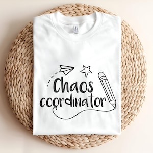 以下が含まれることがあります： Chaos coordinatorという黒い文字がプリントされた白いTシャツ。デザインには、手描き風の紙飛行機、五芒星、そして鉛筆の先から曲線が伸びているグラフィックが含まれています。