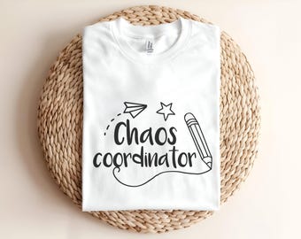 May include: A white t-shirt featuring black text that reads Chaos coordinator. The design includes a hand-drawn style graphic of a paper airplane, a five-pointed star, and a pencil with a curved line extending from the tip of the pencil.
