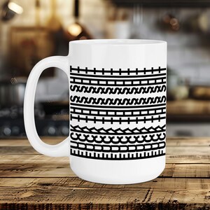 May include: White ceramic mug with a black geometric pattern. The design features horizontal bands of lines, curves, and rectangles. The mug has a large handle and is set on a wooden surface.