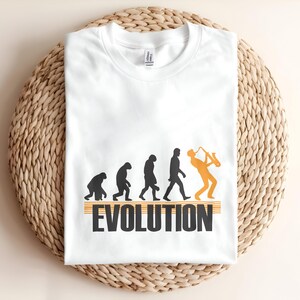 May include: White t-shirt with a graphic depicting the evolution of man, ending with a saxophone player. The word "EVOLUTION" is printed in black with orange stripes. The shirt is folded on a woven mat.