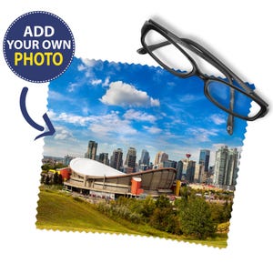 May include: A square microfibre cleaning cloth with pinked edges featuring a printed cityscape of Calgary, including the Saddledome and skyline under a blue sky. A pair of black plastic spectacles rests on the top right corner of the cloth.