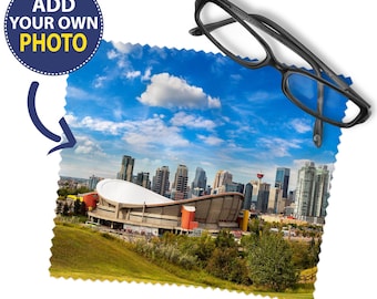 May include: A square microfiber cleaning cloth with pinked edges featuring a printed cityscape of Calgary, including the Saddledome and skyline under a blue sky. A pair of black plastic eyeglasses rests on the top right corner of the cloth.