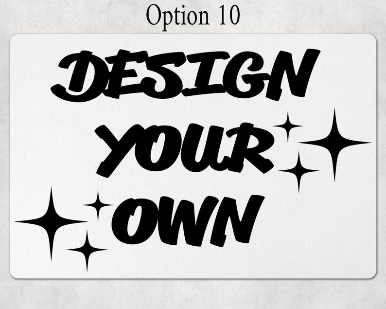 May include: A rectangular white sign featuring the black text "DESIGN YOUR OWN" in a bold, stylized font. The design is accented by several four-pointed star shapes in black, positioned to the left of the bottom word and to the right of the middle word.