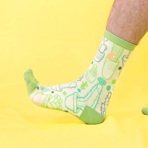 Puede incluir: Un par de calcetines novedosos con un estampado de cactus verde claro y blanco. Los calcetines tienen puños, talones y punteras verdes. El fondo es amarillo brillante.