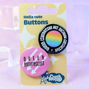 Puede incluir: Dos pines de botón en una tarjeta amarilla con el texto "Hella cute Buttons". El primer pin es negro con un arcoíris y el texto "Queer Liberation Hot Rainbow Capitalism". El segundo pin es rosa con el texto "Queer Anti-Fascist" y dos flechas.