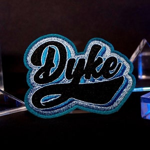 May include: A blue and black embroidered patch with the word "Dyke" in a stylized font. The patch is surrounded by clear glass prisms.