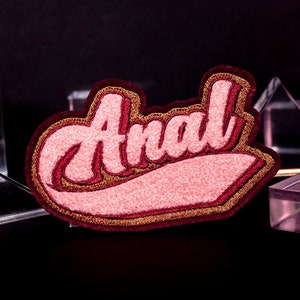 Peut inclure: Un patch brodé rose et or avec le mot "Anal" dans un style de police rétro.