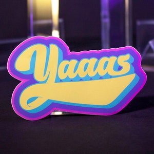 Può includere: Un adesivo con la parola "Yaaas" scritta in un font stilizzato. L'adesivo è giallo, blu e viola.